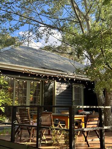 A cozy outdoor deck with wooden chairs and table, string lights above, surrounded by leafy greenery and trees, attached to a quaint house under blue sky.