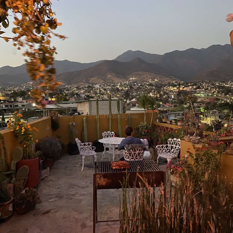 Eine Person sitzt auf einer Dachterrasse mit weißen Stühlen und einem Tisch, umgeben von Pflanzen, und blickt in der Abenddämmerung auf die Stadt und die Berge. Friedliche Atmosphäre.