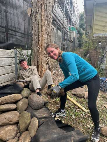 Dos mujeres construyen una pared de roca al aire libre. Una está sentada y sonríe, mientras que la otra, de pie y con guantes, sostiene una pesada piedra, también sonriente.