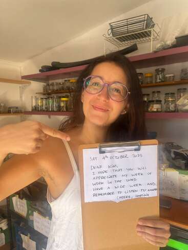 Una mujer con gafas sonríe y señala un portapapeles con una nota manuscrita. Está en un taller lleno de frascos y suministros.