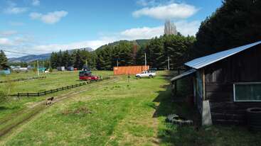 Eine ländliche Szene mit grünem Gras, einem Holzhaus, zwei geparkten Fahrzeugen, einem Hund, einem orangefarbenen Frachtcontainer, Kiefern, blauem Himmel und fernen Bergen.