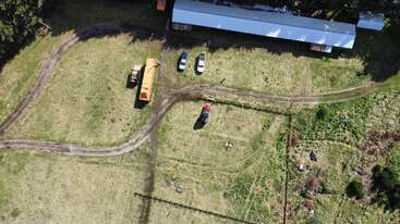 Dieses Luftbild zeigt ein grasbewachsenes Feld mit einer unbefestigten Straße, einem großen Schuppen, geparkten Autos, Baumaschinen und einigen Bäumen und Pflanzen in der Umgebung.
