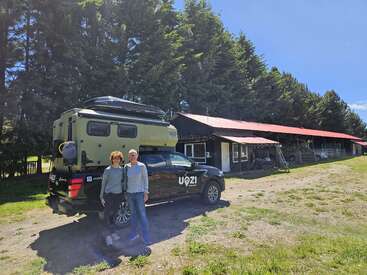 Ein lächelndes Paar steht neben einem schwarzen Geländewagen mit der Aufschrift "VOZI". Hinter ihnen steht ein rustikales Gebäude, umgeben von hohen Kiefern unter einem sonnigen Himmel.
