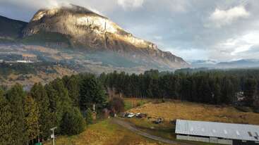 Ein majestätischer Berg erhebt sich hinter einem dichten Wald, der in goldenes Sonnenlicht getaucht ist. Unten liegt eine ländliche Landschaft mit verstreuten Bäumen, ein paar Fahrzeugen und einem großen Gebäude.