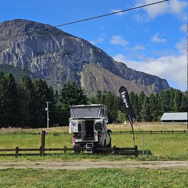 A camper vehicle is parked on green grass with a raised tent roof. In the background, a rugged mountain rises above dense trees under a blue sky.