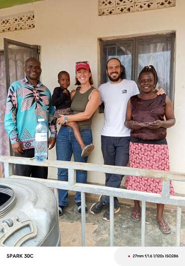 Five people, including a child, stand smiling together outside a house. A water bottle sits on a railing in front. The atmosphere is warm and friendly.