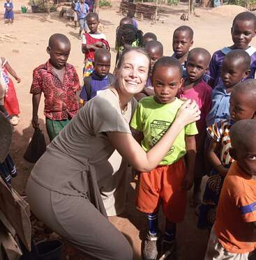 Eine lächelnde Frau umarmt einen kleinen Jungen in einem grünen Hemd, umgeben von Kindern in bunten Kleidern im Freien, die eine fröhliche und lebhafte Gruppenszene bilden.