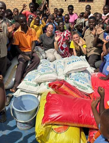 Eine fröhliche Gruppe von Kindern und Erwachsenen versammelt sich lächelnd und winkend um Lebensmittelvorräte, während im Vordergrund Säcke mit Getreide und Hilfsgütern zu sehen sind.
