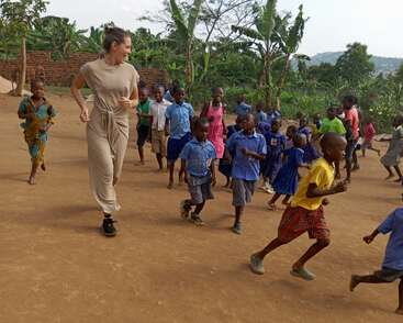 Eine lächelnde Frau in Beige läuft fröhlich mit einer großen Gruppe von Schulkindern im Freien. Die Kinder tragen bunte Uniformen, wirken fröhlich und energiegeladen und sind von Grün umgeben.