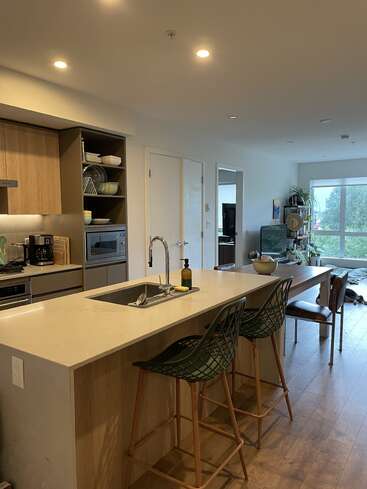 This image shows a modern kitchen with a marble island, two barstools, wood cabinets, open shelving, sink, appliances, and adjoining dining and living areas with large windows.
