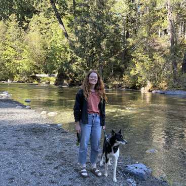 A woman with long red hair stands smiling by a clear river, holding a black and white dog on a leash. Lush green forest surrounds them.