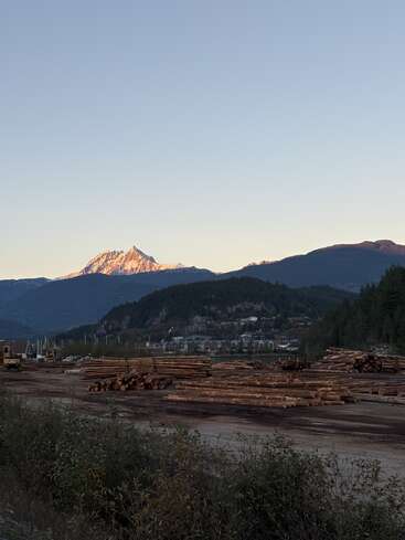A serene mountain landscape at dusk, with stacks of logs in the foreground. Snow-capped peak glows with sunset light, small town nestled amid trees below.