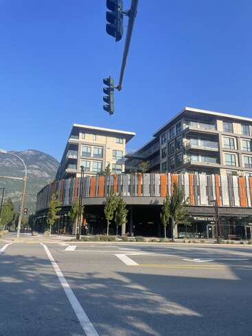 A modern apartment building with colorful vertical panels sits on a street corner beneath clear blue skies, mountains in the background, and a traffic light overhead.