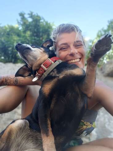 A smiling person with short blonde hair enjoys a joyful moment outdoors, holding a happy black and brown dog with a red collar and playful posture.