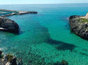 Crystal clear turquoise water surrounded by rocky cliffs, sunlit and inviting. A small pier extends into the sparkling sea, creating a peaceful coastal scene.