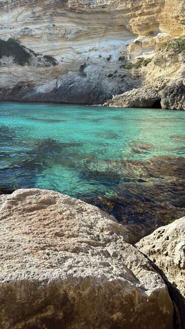 Crystal-clear turquoise water bordered by large, weathered rocks and dramatic, layered cliffs. Sunlight illuminates the serene cove, creating a peaceful and inviting natural setting.