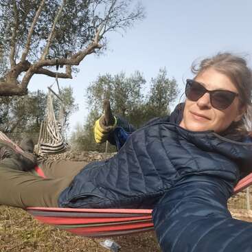 A woman wearing sunglasses and gloves relaxes on a striped hammock outdoors, giving a thumbs up. Trees and nature surround her, and she looks content.