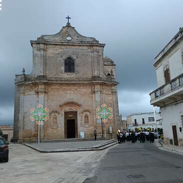 An old stone church stands under a cloudy sky. Colorful illuminated decorations and a marching band with brass instruments gather in front on the quiet street.