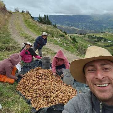 Un hombre sonriente se hace un selfie con un grupo de personas que cosechan patatas al aire libre en la ladera de una montaña, con unas pintorescas colinas verdes y el cielo nublado de fondo.