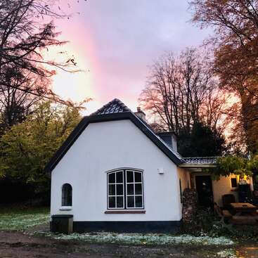A charming white cottage stands amidst trees with autumn foliage. The sky glows pink and orange, creating a serene, peaceful atmosphere as day turns to dusk.