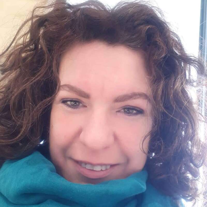 A woman with curly brown hair smiles at the camera. She wears a teal scarf and is positioned in bright, natural light, creating a cheerful close-up portrait.