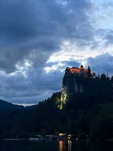 Un majestueux château illuminé se dresse au sommet d'une falaise abrupte, surplombant un sombre paysage forestier. Le ciel nuageux dramatique contraste avec les eaux paisibles en contrebas au crépuscule.