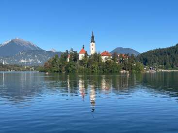 Un lac serein reflète une église pittoresque avec un haut clocher, entourée d'arbres luxuriants, de montagnes et sous un ciel bleu clair, créant une beauté tranquille.