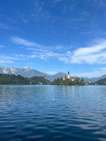 Un lac serein se reflète dans le ciel bleu, avec une petite île-église au centre, entourée de montagnes et de forêts sous des nuages vaporeux et le soleil.