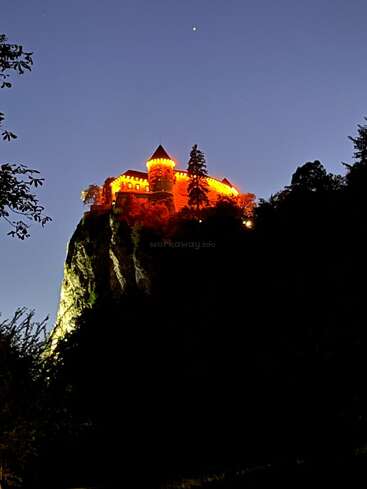 Un château illuminé se trouve au sommet d'une falaise abrupte, brillant en orange dans le ciel du crépuscule, entouré d'arbres et se découpant sur l'obscurité qui s'approche.