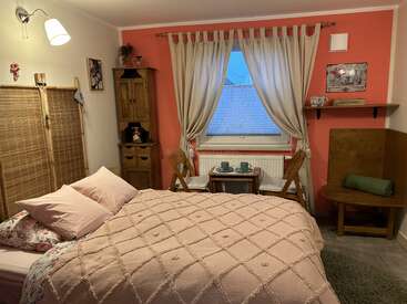 Une chambre à coucher confortable avec un mur d'accent rose comprend un lit, des meubles en bois, une petite table avec deux chaises, des rideaux et un éclairage d'ambiance chaleureux. Atmosphère accueillante.