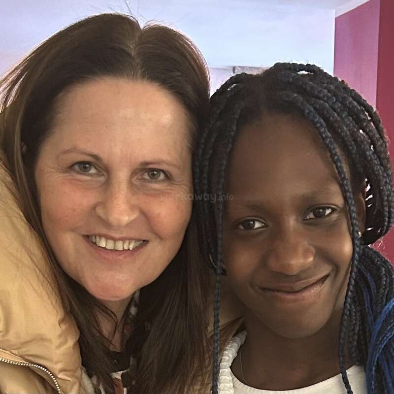 Two women, one with straight brown hair and one with braided blue hair, smile warmly together indoors against a colorful background, showing friendship and joy.