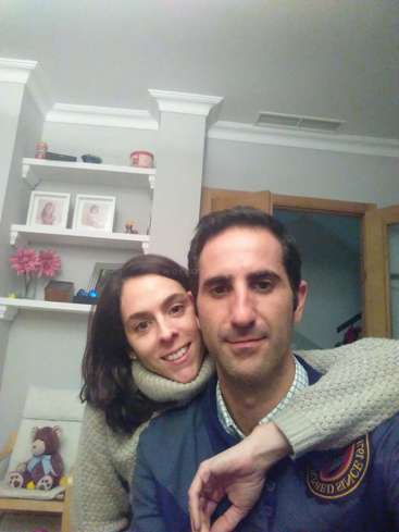 Das Bild zeigt einen Mann und eine Frau, die für ein Selfie in einem gemütlichen Zimmer posieren, wobei die Frau den Mann von hinten umarmt.