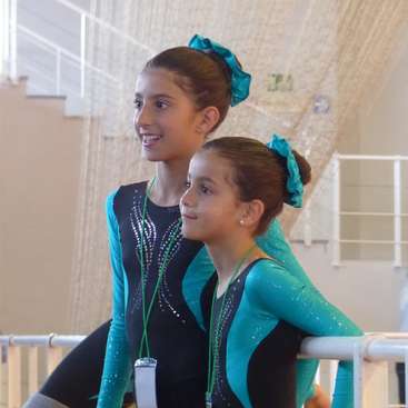 The image shows two young girls in matching gymnastics leotards, standing together in front of a white railing, with a watermark across the center.
