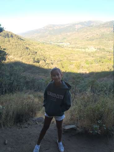 Das Bild zeigt ein junges Mädchen, das auf einem Feldweg steht, umgeben von üppigem Grün und Bergen im Hintergrund, mit einer ruhigen und natürlichen Atmosphäre.