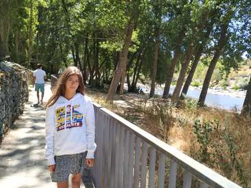 Das Bild zeigt ein junges Mädchen, das auf einer Brücke steht und einen weißen Kapuzenpulli mit einem Logo trägt, mit Bäumen und einem Fluss im Hintergrund und einem Mann, der hinter ihr geht.