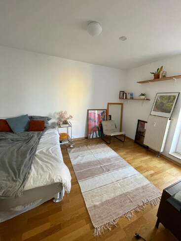 The image depicts a cozy bedroom with a bed, a rug, and a chair, featuring white walls, a wooden floor, and a few decorative elements, including a mirror and a painting.