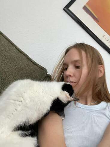 The image depicts a young woman with blonde hair, wearing a white shirt, sitting on a couch with a black and white cat leaning against her.