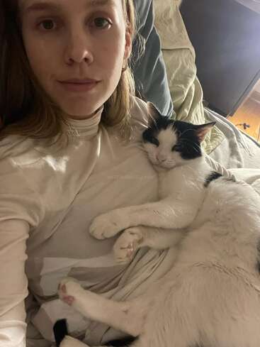 The image shows a woman with blonde hair and a white shirt holding a black and white cat on her chest. The cat is looking at the camera.