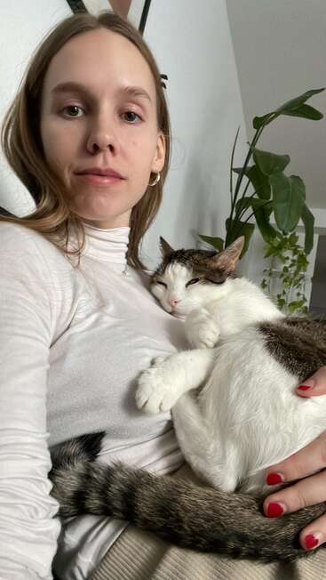 The image shows a young woman with long brown hair, wearing a white turtleneck, holding a cat with white and brown fur, in a room with a plant in the background.