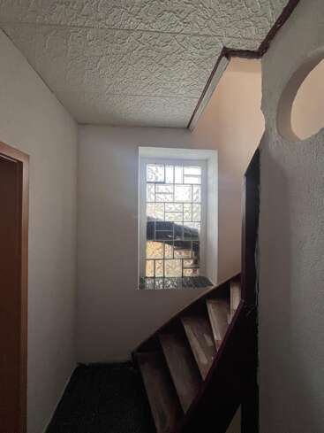 Un escalier étroit et faiblement éclairé mène à une fenêtre en briques de verre, entourée de murs et d'un plafond blancs texturés, créant ainsi un espace intérieur fermé et inachevé.