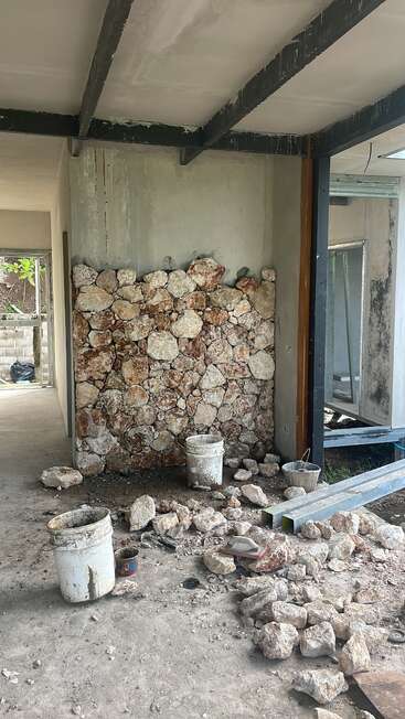 Há uma parede de pedra parcialmente construída dentro de um prédio inacabado. Baldes, pedras e ferramentas de construção estão espalhados pelo chão empoeirado. Vêem-se vigas expostas e paredes nuas.