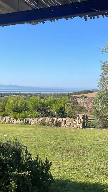 Um jardim tranquilo, com grama verdejante, um muro de pedra e arbustos, tem vista para colinas distantes, pinheiros e um mar azul e calmo sob um céu limpo.