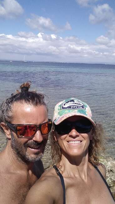 Um casal sorridente posa para uma selfie à beira do mar azul cristalino, ambos usando óculos escuros. A mulher usa um chapéu com a inscrição “Costa Rica”. Barcos à vela ao fundo.