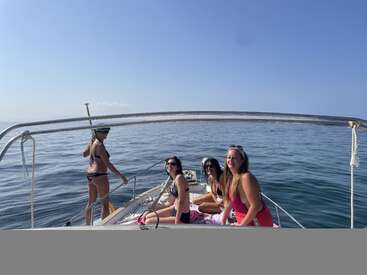 Quatro mulheres relaxam em um barco no mar aberto, aproveitando o dia ensolarado. Três estão sentadas, uma está em pé com um remo, todas sorridentes e felizes.