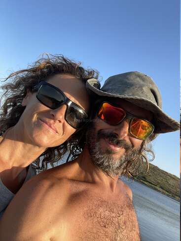 Um casal sorridente posa para uma selfie ao ar livre, ambos usando óculos escuros. O homem usa chapéu e tem barba. O céu azul brilhante e a água ficam atrás deles.