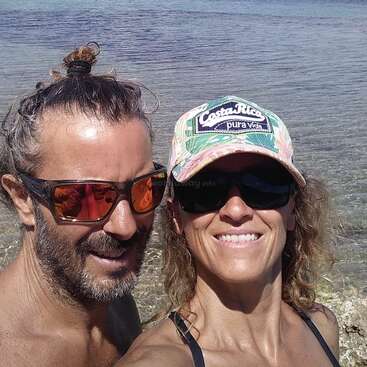 Un couple souriant pose pour un selfie au bord d'une mer d'un bleu limpide, tous deux portant des lunettes de soleil. La femme porte une casquette « Costa Rica ». Des voiliers sont visibles à l'arrière-plan.