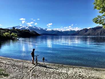 Das Bild zeigt eine ruhige Seeszene mit einer Person, die am Ufer steht, einem Fahrrad in der Nähe und einem Kind, das im Wasser spielt, vor einer Bergkulisse.