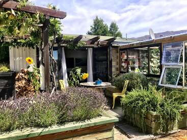 Das Bild zeigt einen ruhigen Terrassenbereich mit einer hölzernen Pergola, einem Tisch mit Stühlen und üppigem Grün vor einem wolkenverhangenen Hintergrund.