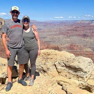Das Bild zeigt einen Mann und eine Frau, die auf einem Felsen stehen, hinter ihnen erstreckt sich eine weite Canyonlandschaft unter einem klaren blauen Himmel.