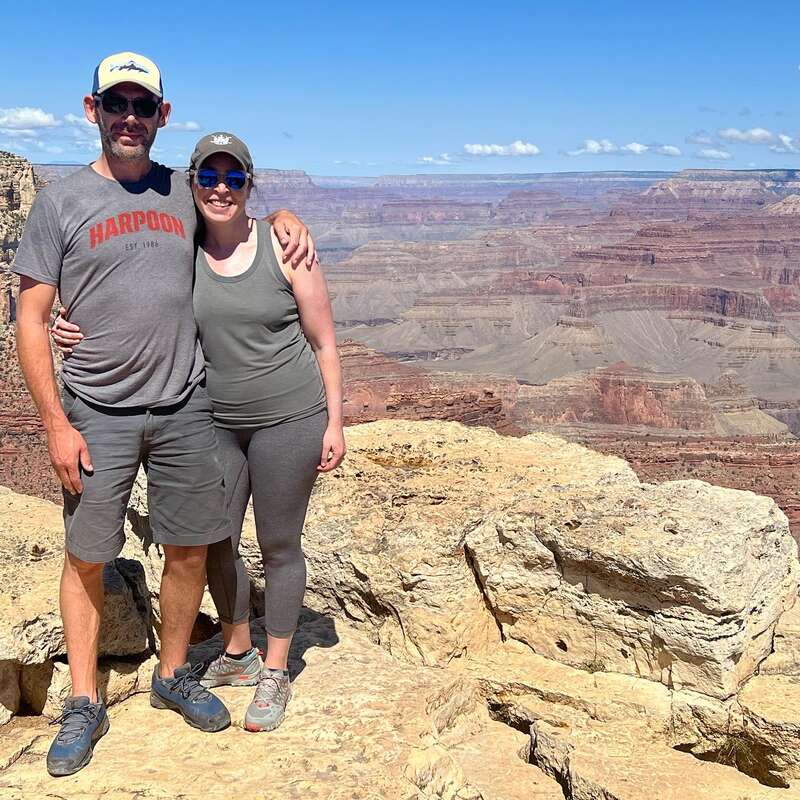 Das Bild zeigt einen Mann und eine Frau, die auf einem Felsen stehen, hinter ihnen erstreckt sich eine weite Canyonlandschaft unter einem klaren blauen Himmel.